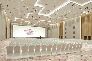 Ein Konferenzraum mit Reihen weißer Stühle, einem großen Bildschirm, der Text anzeigt, und einem modernen Deckenbau.