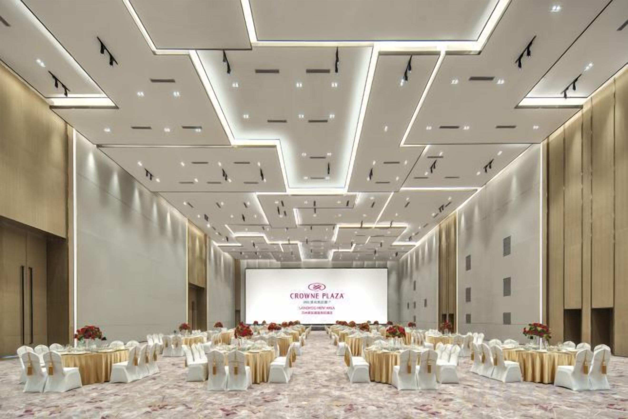 A conference room with a stage, rows of tables with chairs, and a large screen displaying 'CROWNE PLAZA'.