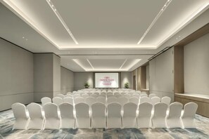 Une salle de conférence avec des rangées de chaises blanches, un pupitre et un grand écran affichant du texte.