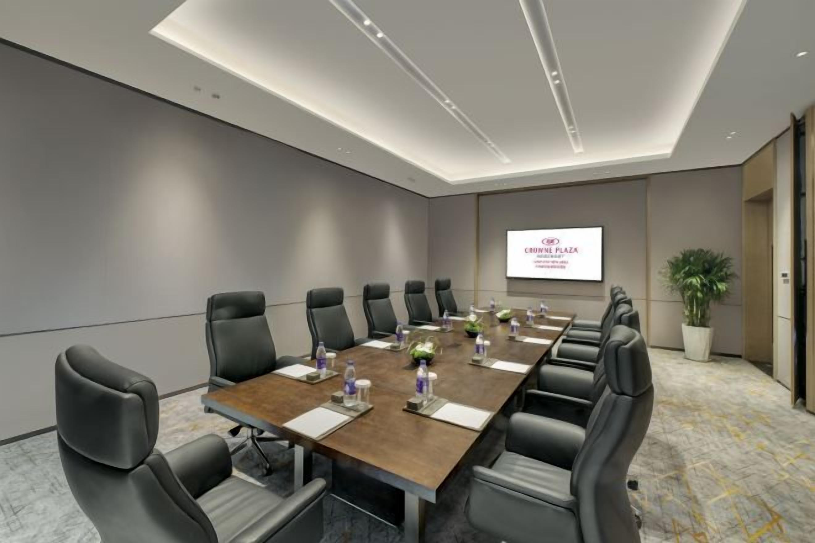 A conference room with a long table, multiple chairs, a projector screen, and a potted plant.
