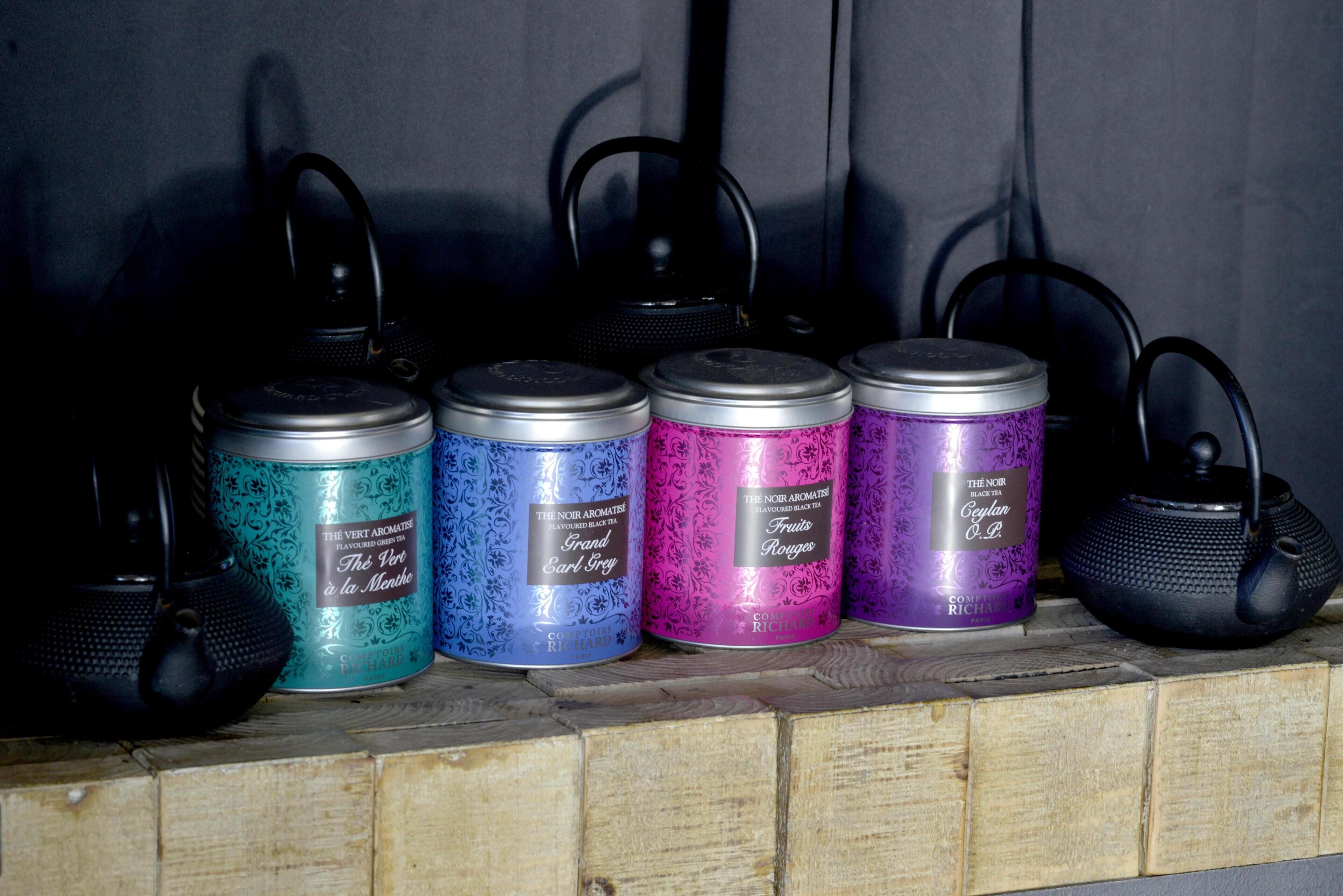 Four tea kettles with lids, each in a different color, placed on a wooden shelf.