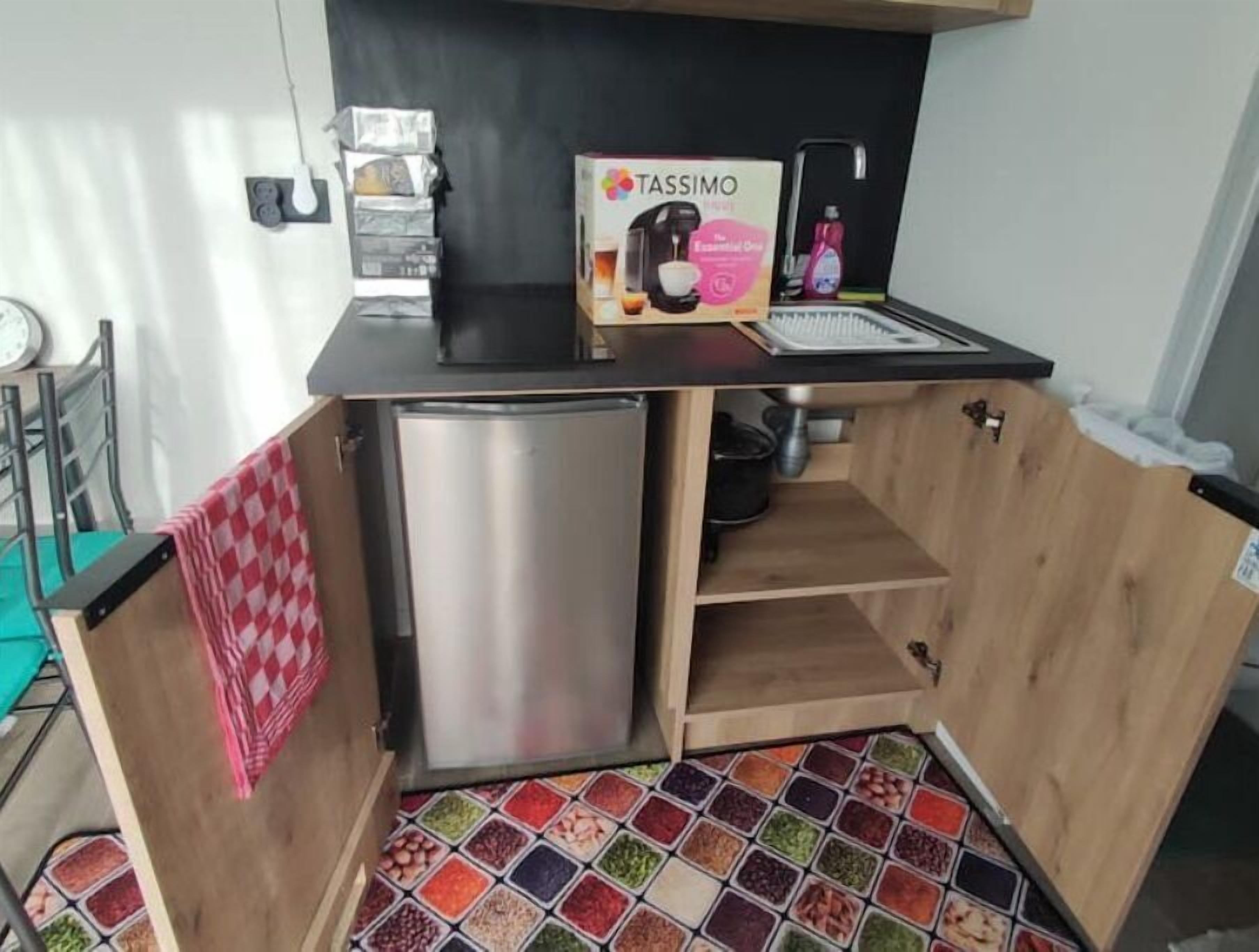 A compact kitchen with a stainless steel mini-fridge, a Tassimo coffee maker, and a colorful spice rack.