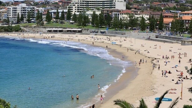 Ein Strand mit badenden Personen und Sonnenanbetern, einem Rettungsturm und Gebäuden im Hintergrund.