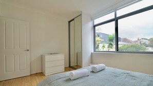 Ein Schlafzimmer mit einem Bett, einer weißen Kommode, einem großen Fenster mit Blick auf Gebäude und Bäume sowie einer Tür.