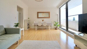 Ein modernes Wohnzimmer mit einer Couch, einem Esstisch mit Stühlen, einem Fernseher und einem großen Fenster.