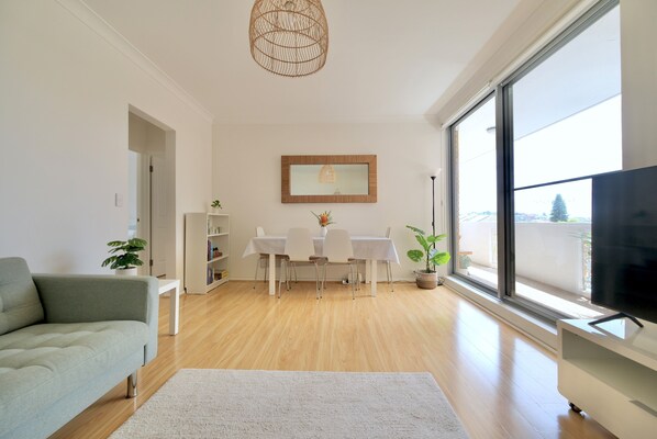 A modern living room with a sofa, a dining table with chairs, a TV, and a large window. - Modern Poolside Coogee Beach Sanctuary (Sydney)