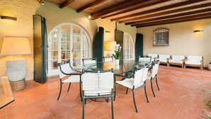 Uma área de refeições com mesa de vidro e cadeiras, cercada por janelas em arco e piso ladrilhado.