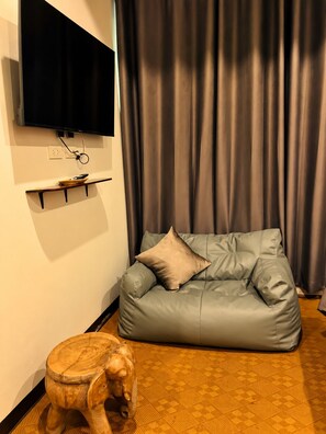 A living room with a flat-screen TV mounted on the wall, a grey sofa with a cushion, a small wooden stool, and a patterned carpet. - CHERN CHIANGMAI BOUTIQUE (Chiang Mai)