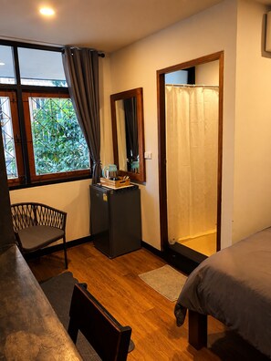 A compact room with a bed, a small desk, a chair, a window with curtains, a mirror, and a shower area. - CHERN CHIANGMAI BOUTIQUE (Chiang Mai)