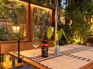 A table with a bottle of wine, a glass, and a plant in a vase, set against a window with a view of greenery. - CHERN CHIANGMAI BOUTIQUE (Chiang Mai)