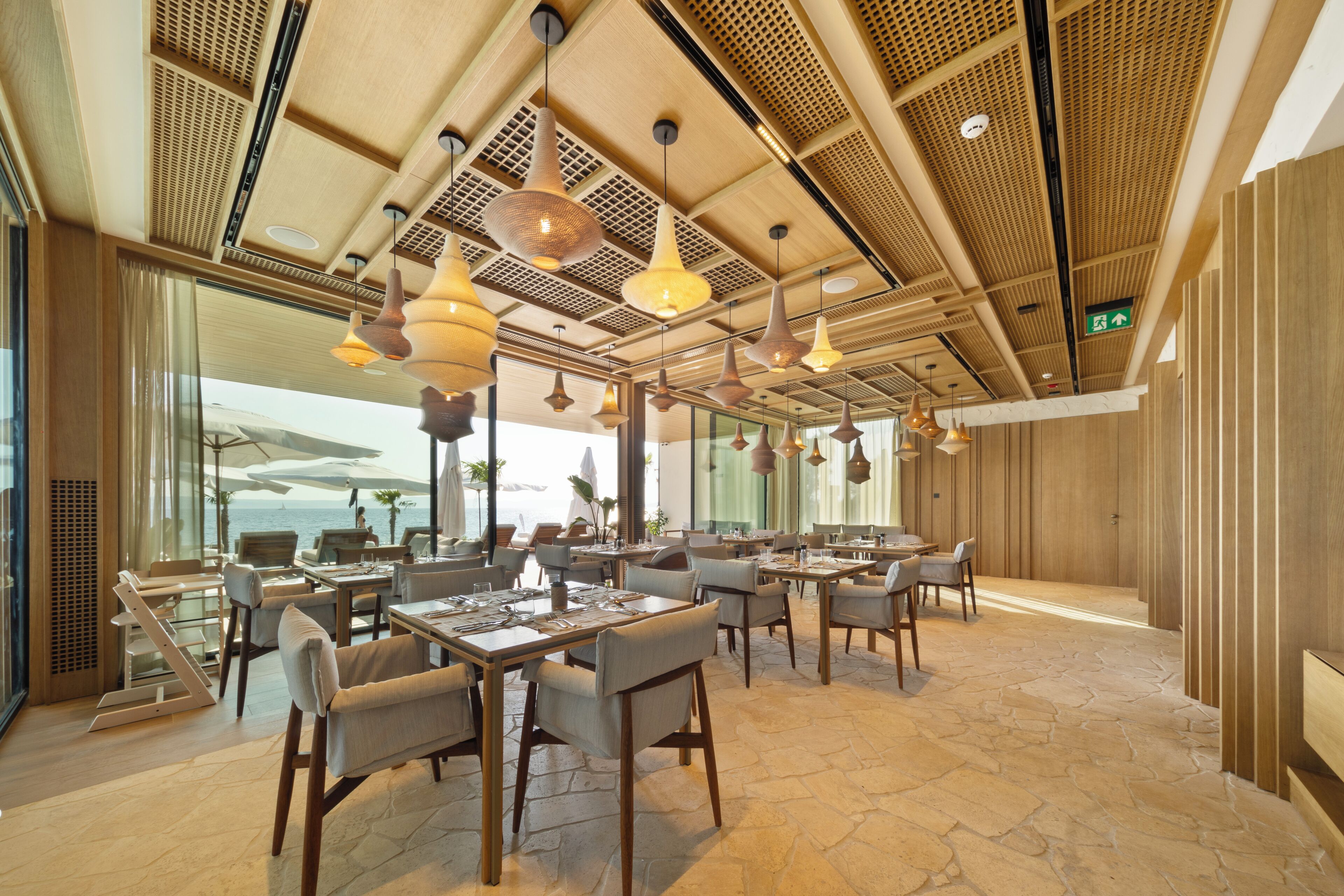Un'ampia sala ristorante con numerosi tavoli e sedie, ampie finestre che offrono vista sul mare e un design moderno del soffitto con lampade pendenti.