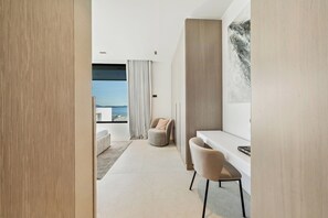 A modern hotel room with a minimalist design, featuring a large window with a view of the sea, a comfortable seating area, and a small desk.