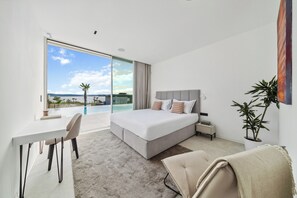 A modern bedroom with a large bed, a desk, and a chair. It features a sliding glass door leading to a balcony with a view of the sea.