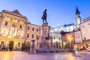 Una plaza pública con una estatua, edificios de estilo clásico y una torre del reloj.