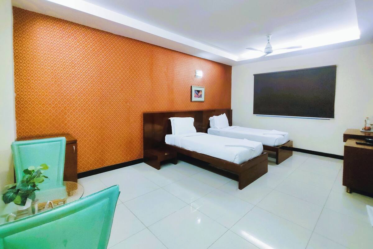 Quarto de hotel com duas camas de solteiro, uma escrivaninha, uma cadeira, uma pequena mesa com uma planta e uma janela grande.
