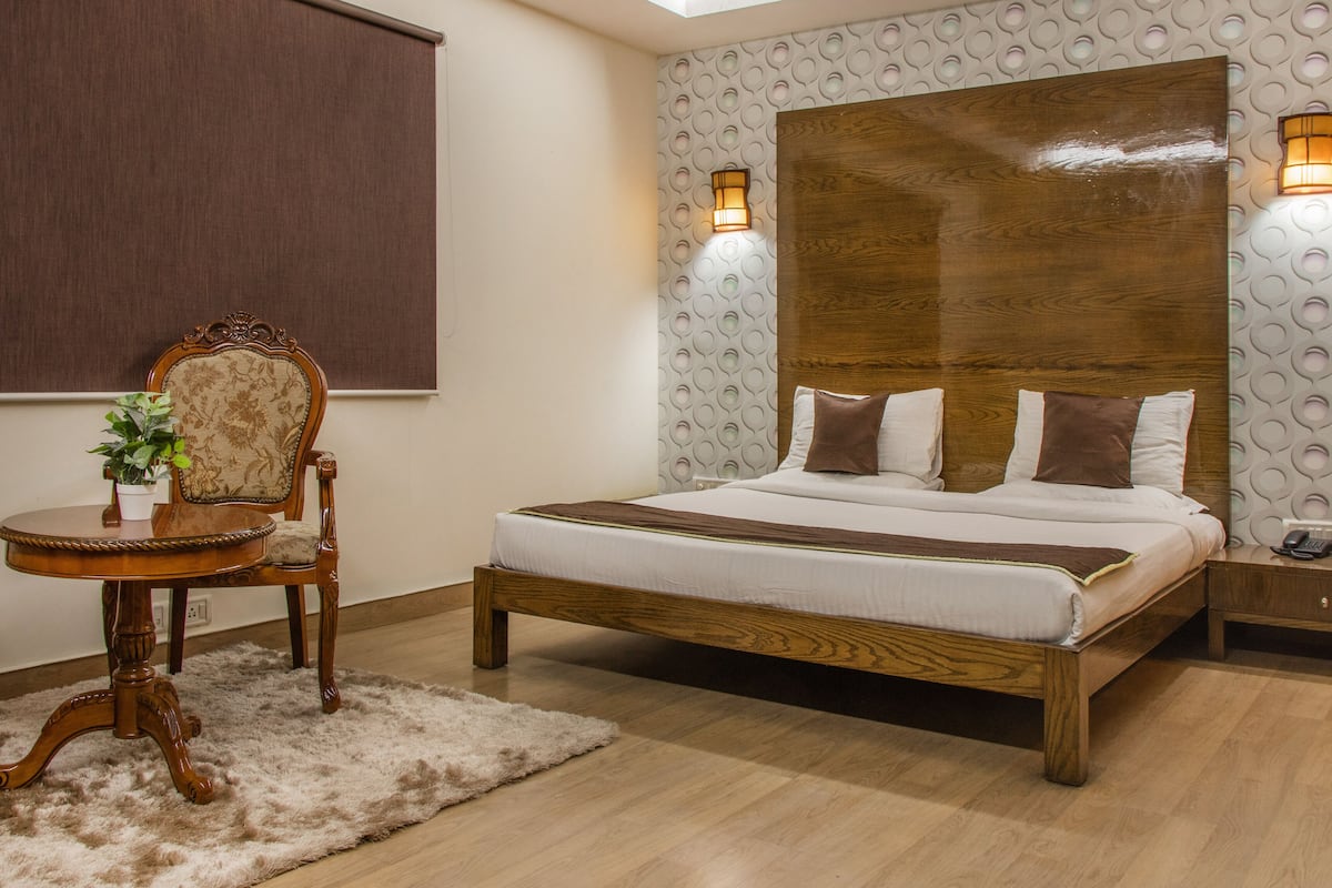Quarto bem arrumado com uma cama de madeira, uma pequena mesa redonda com uma planta, uma cadeira e uma mesa de cabeceira com um telefone.