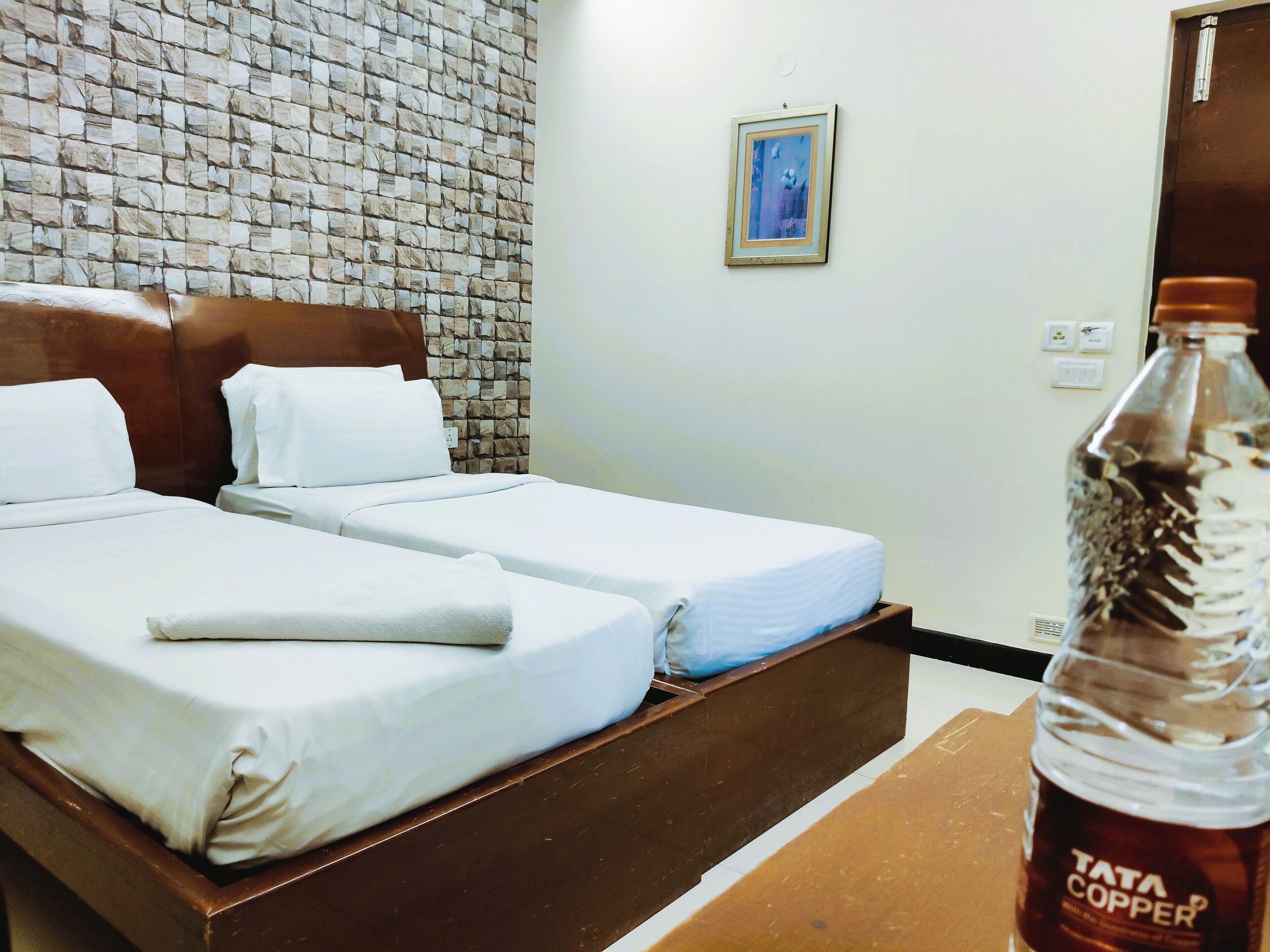 A hotel room with a brick-patterned wall, two beds with white linens, a framed picture on the wall, and a bottle of Tata Copper on the table.