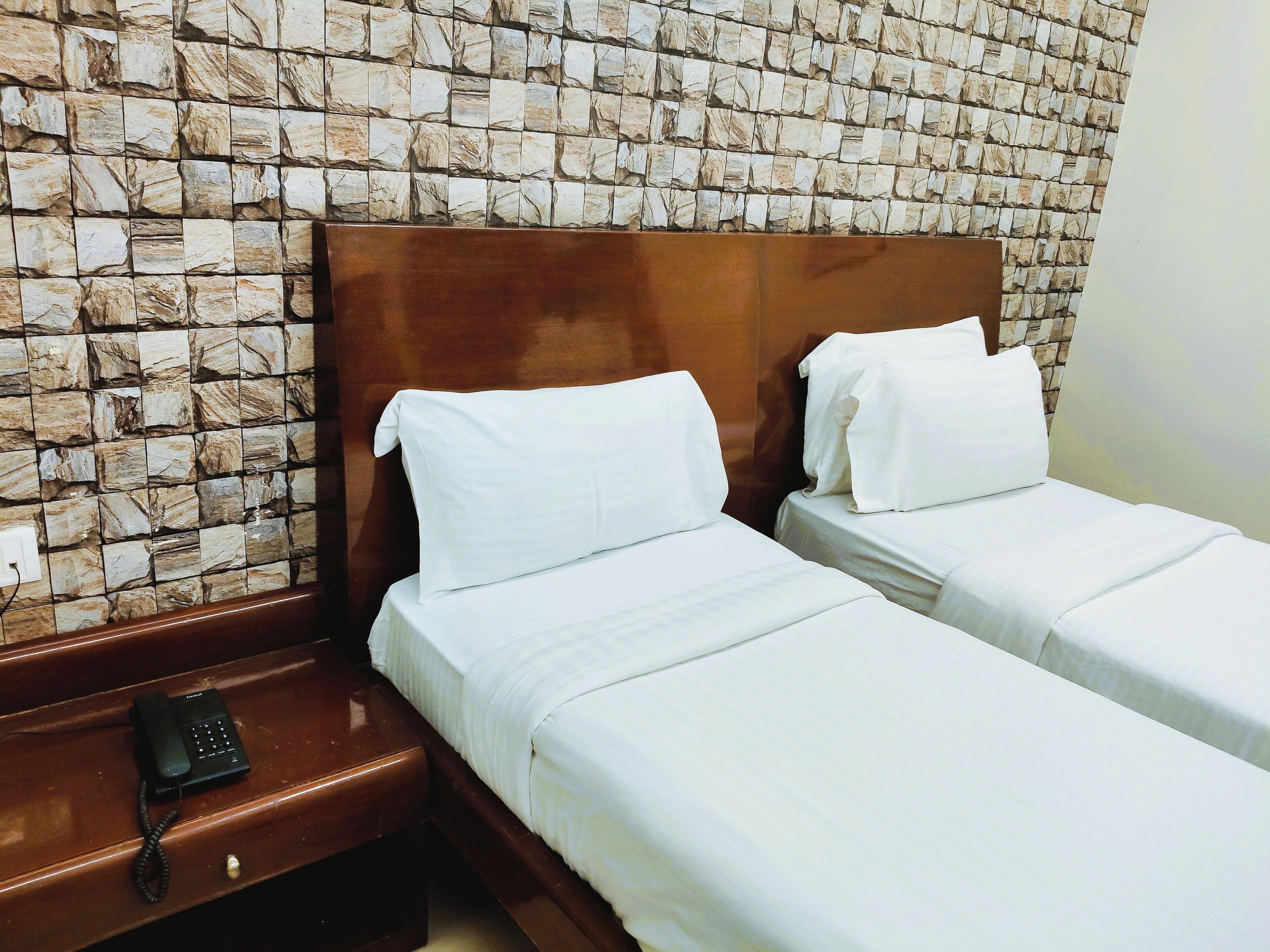 A hotel room with two single beds, a wooden headboard, a nightstand with a telephone, and a stone-patterned wall.