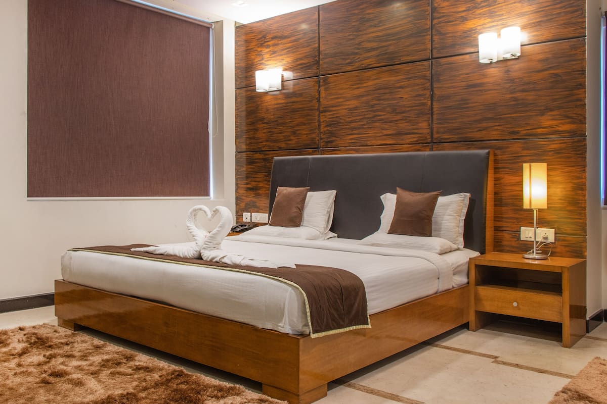 Quarto de hotel moderno com cabeceira de madeira, cama grande com roupa de cama branca e castanha, tapete marrom fofinho e mesa de cabeceira com abajur.