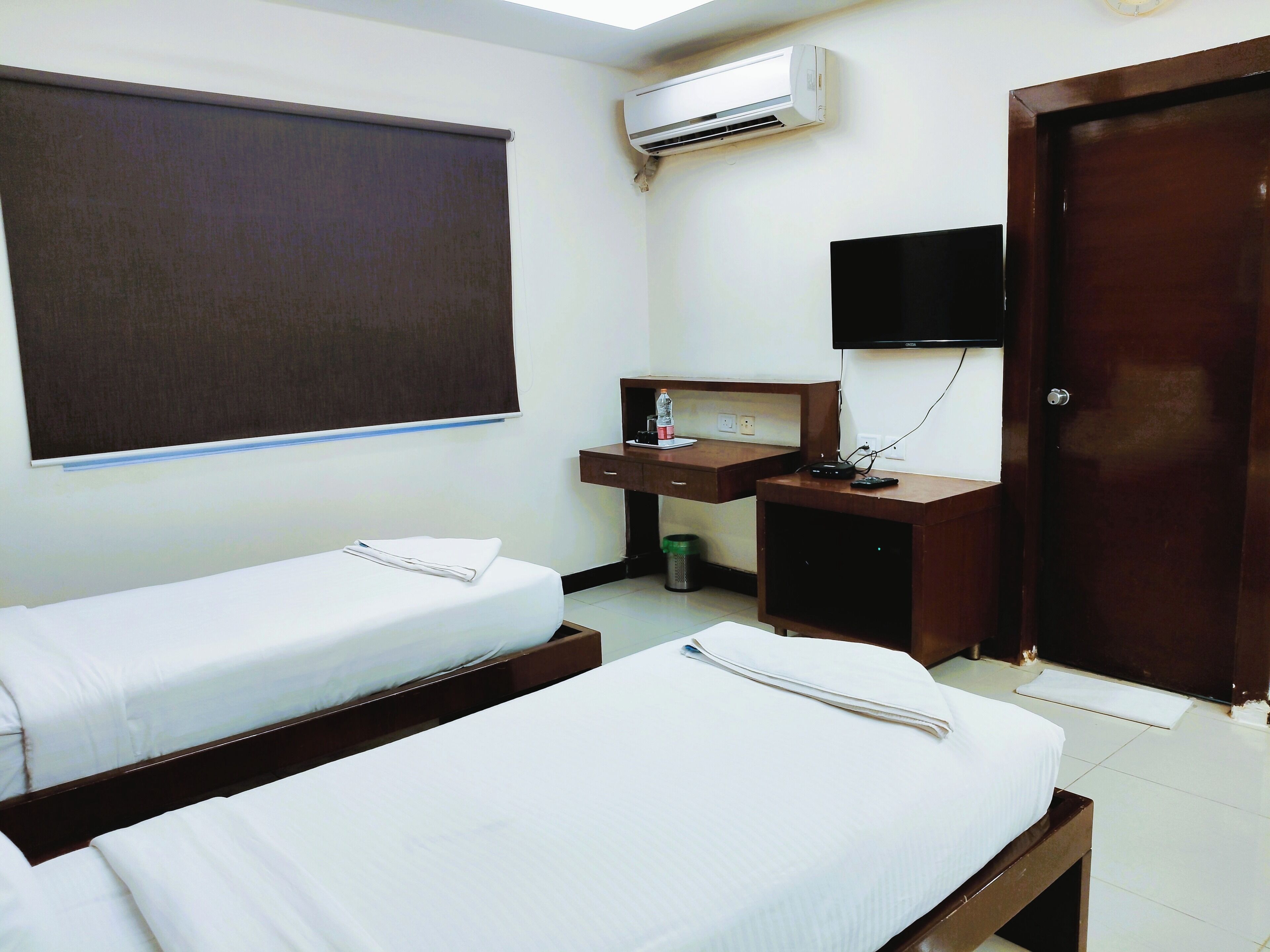 A hotel room with two single beds, a desk with a chair, a TV, and an air conditioning unit.