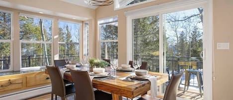 A dining area with a wooden table set for four, surrounded by chairs, large windows, and a view of trees.