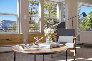 Uma sala de estar moderna com uma mesa de madeira redonda, uma poltrona confortável e um sofá. Há uma janela grande que oferece vista para árvores e montanhas.