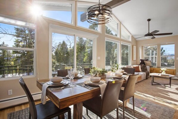 A dining area with a wooden table set for six, surrounded by chairs. Large windows provide a view of trees and a balcony.