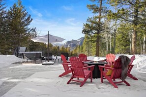 A snow-covered outdoor seating area with red Adirondack chairs and a table, surrounded by trees and mountains in the background.