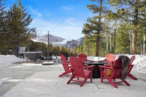 Un'area salotto esterna innevata con sedie Adirondack rosse e un tavolo, circondata da alberi e montagne sullo sfondo.