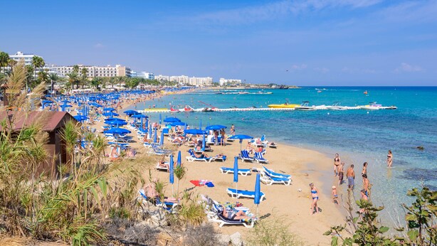 A beach with numerous blue umbrellas and sun loungers, a clear blue sea, and buildings in the background. - Villa Artemis (Paralimni)