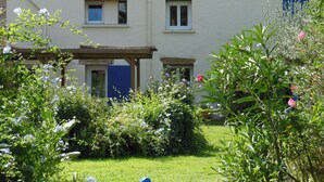 Ein zweistöckiges Gebäude mit Ziegeldach, einer Veranda und einem gepflegten Garten mit verschiedenen Pflanzen und Blumen.
