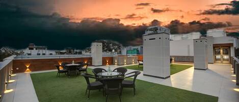 Une terrasse sur le toit, avec une table et des chaises, entourée de gazon synthétique, d’où l’on peut admirer un panorama sur la ville, avec ses bâtiments et un ciel au coucher du soleil.