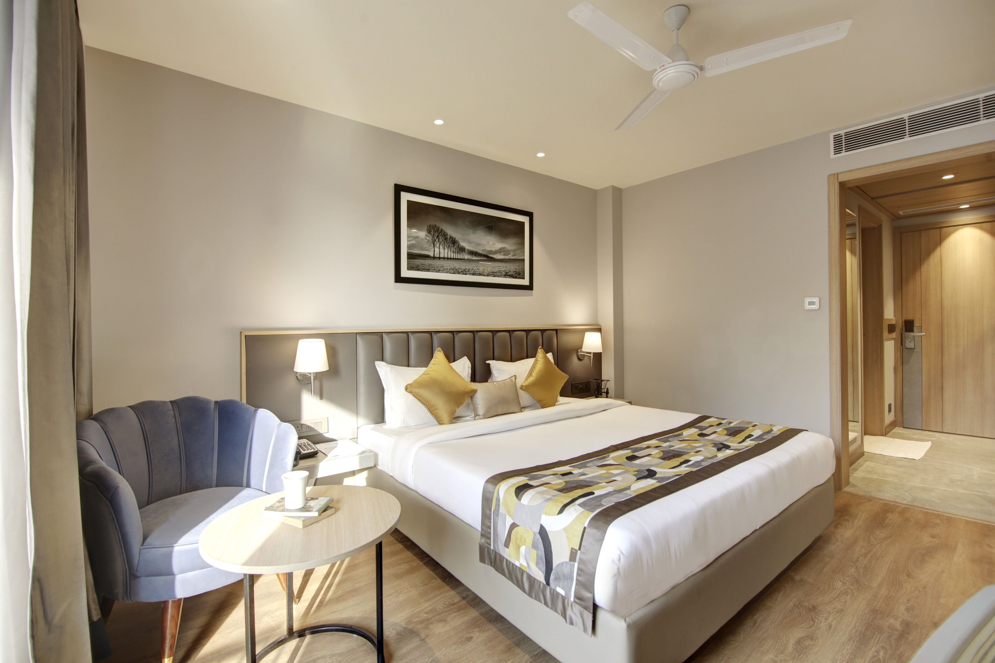A hotel room with a bed, a chair, a small table, a ceiling fan, and a framed picture on the wall.