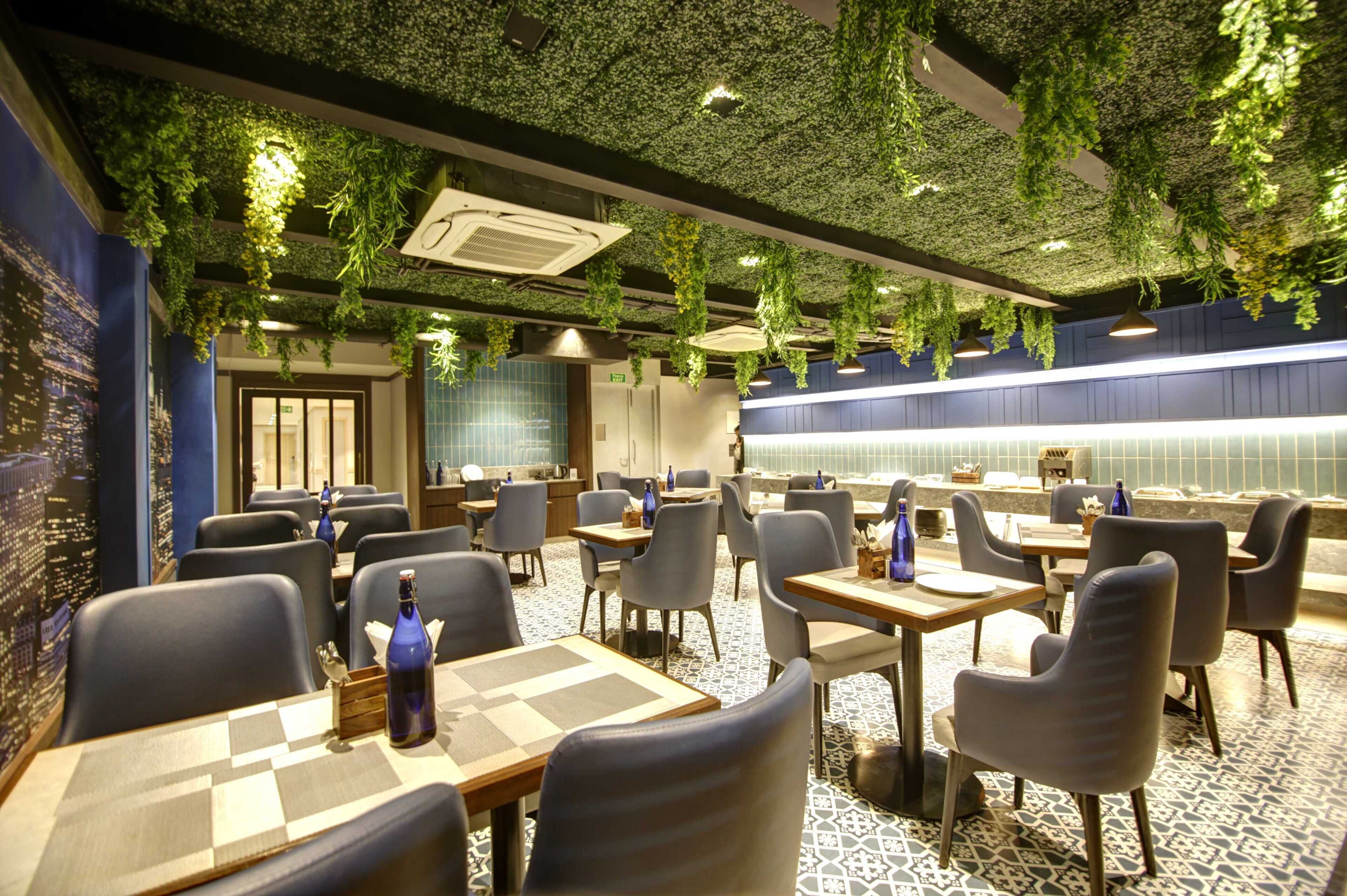 A restaurant with indoor plants hanging from the ceiling, tables with chairs, and a cityscape mural on the wall.