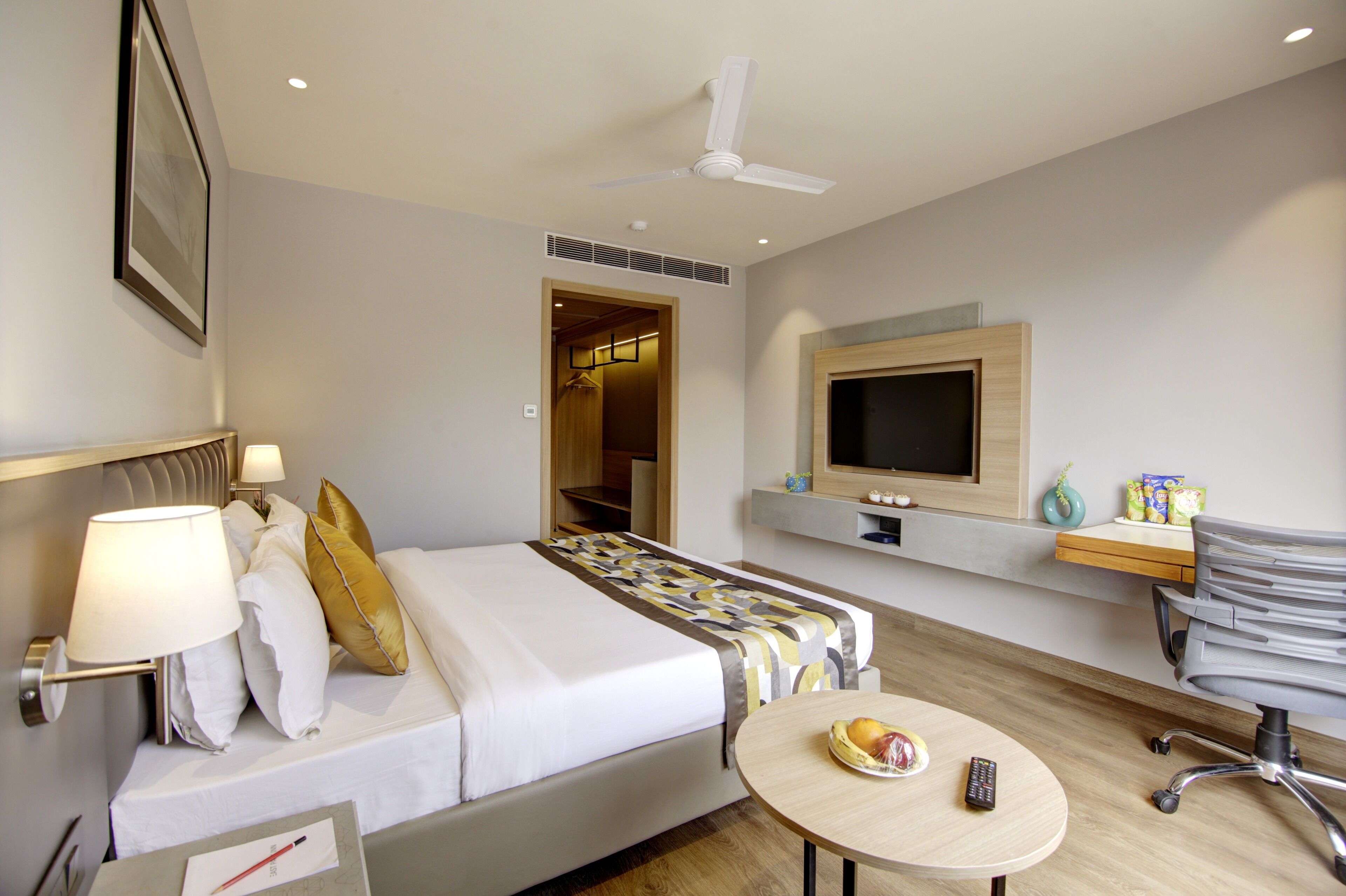 Quarto de hotel com cama, mesa de cabeceira, luminária, televisão e escrivaninha com cadeira.