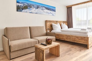 A modern hotel room with a sofa, a bed with wooden headboard, a small wooden side table, and a painting of a snowy mountain landscape.