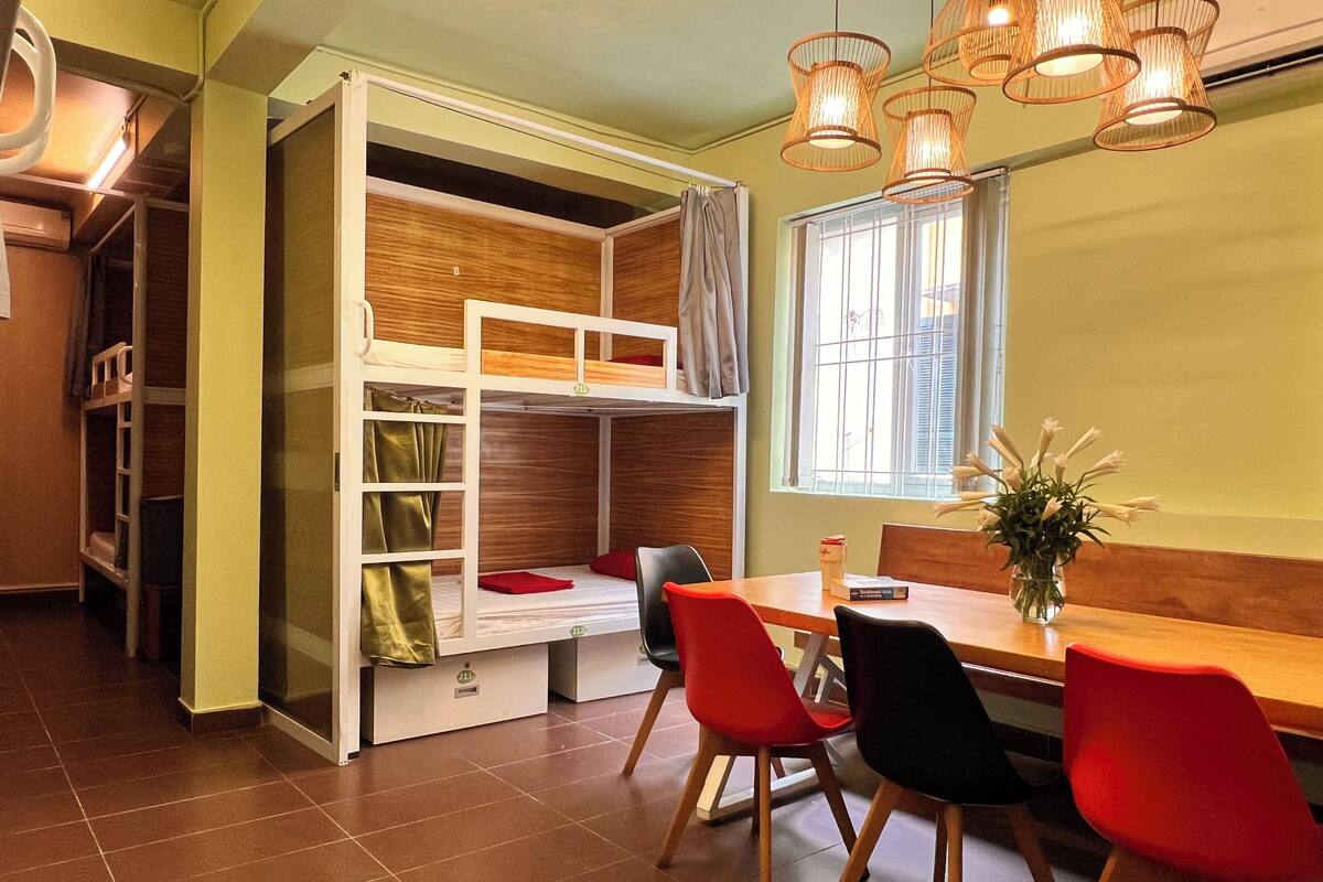 A bunk bed with a desk and chairs, a window with curtains, and a vase with flowers.