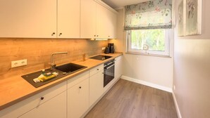 Une cuisine compacte avec des meubles blancs, un plan de travail en bois, un four encastré et une fenêtre agrémentée de rideaux à motifs floraux.