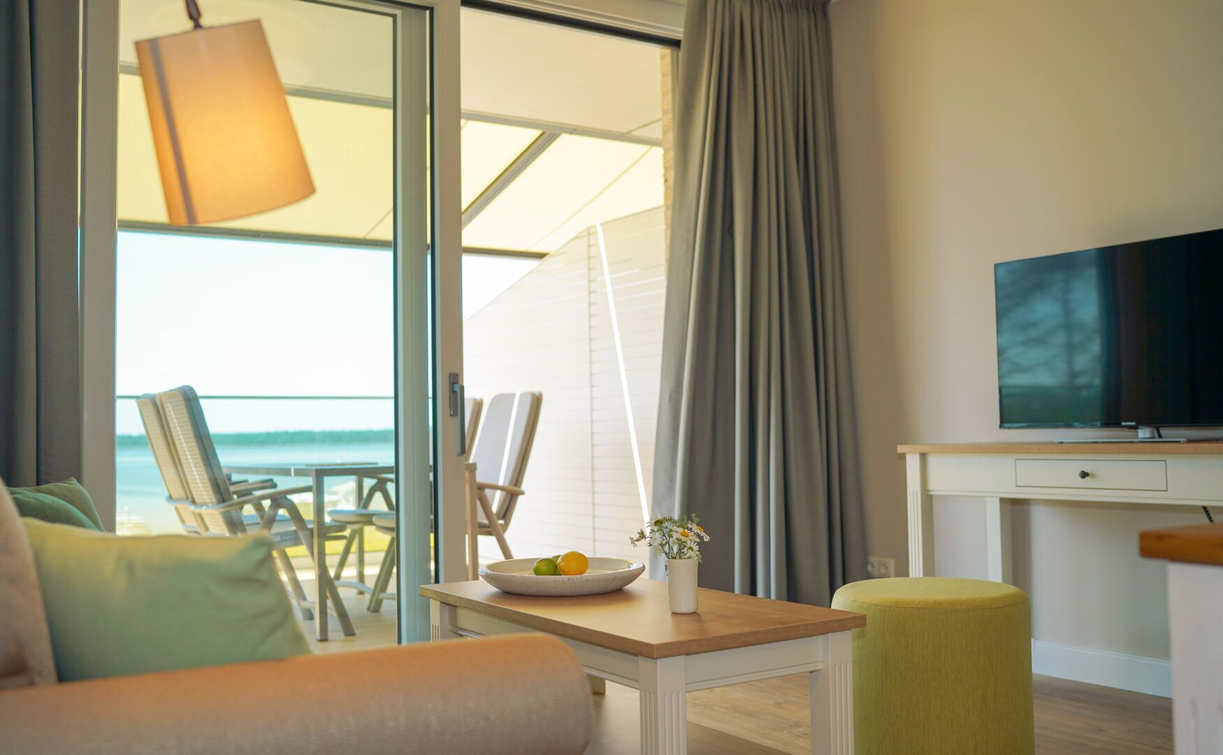 A hotel room with a balcony, a television, a dining table with a bowl of fruit, and a sofa.