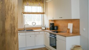 A compact kitchen with white cabinets, a wooden countertop, a built-in oven, a sink, and a window with floral curtains. - Comfortable Upper-Floor Suite with Terrace – Ideal for up to 3 People (Rankwitz)