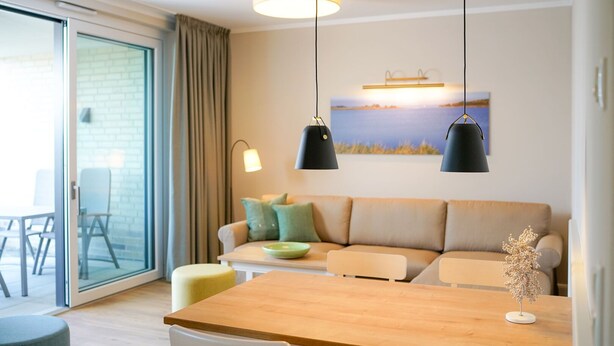 A modern living room with a wooden dining table, white chairs, a beige sofa with green cushions, and two pendant lights. - Comfortable Upper-Floor Suite with Terrace – Ideal for up to 3 People (Rankwitz)