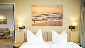 A hotel room with a bed, two bedside lamps, a painting of a beach scene, and a view of a beach through the window. - Comfortable Upper-Floor Suite with Terrace – Ideal for up to 3 People (Rankwitz)