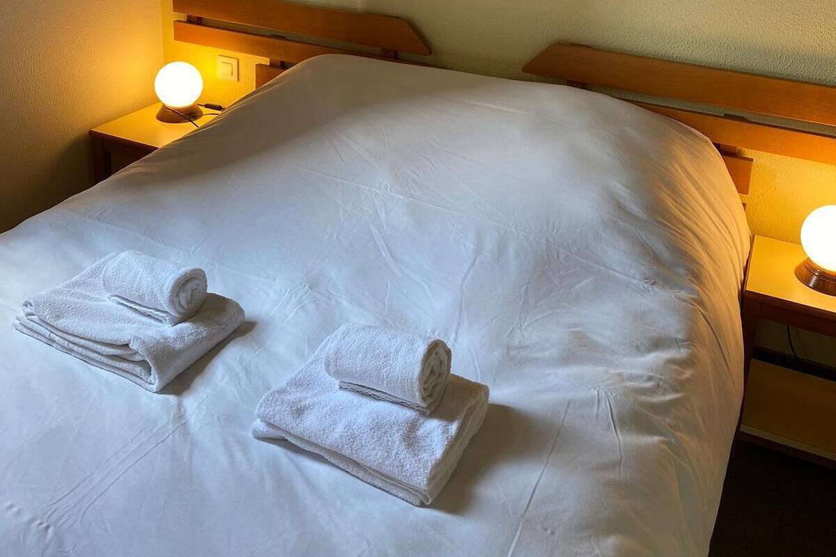A neatly made bed with white linens and two rolled towels, wooden headboard, and bedside lamps.