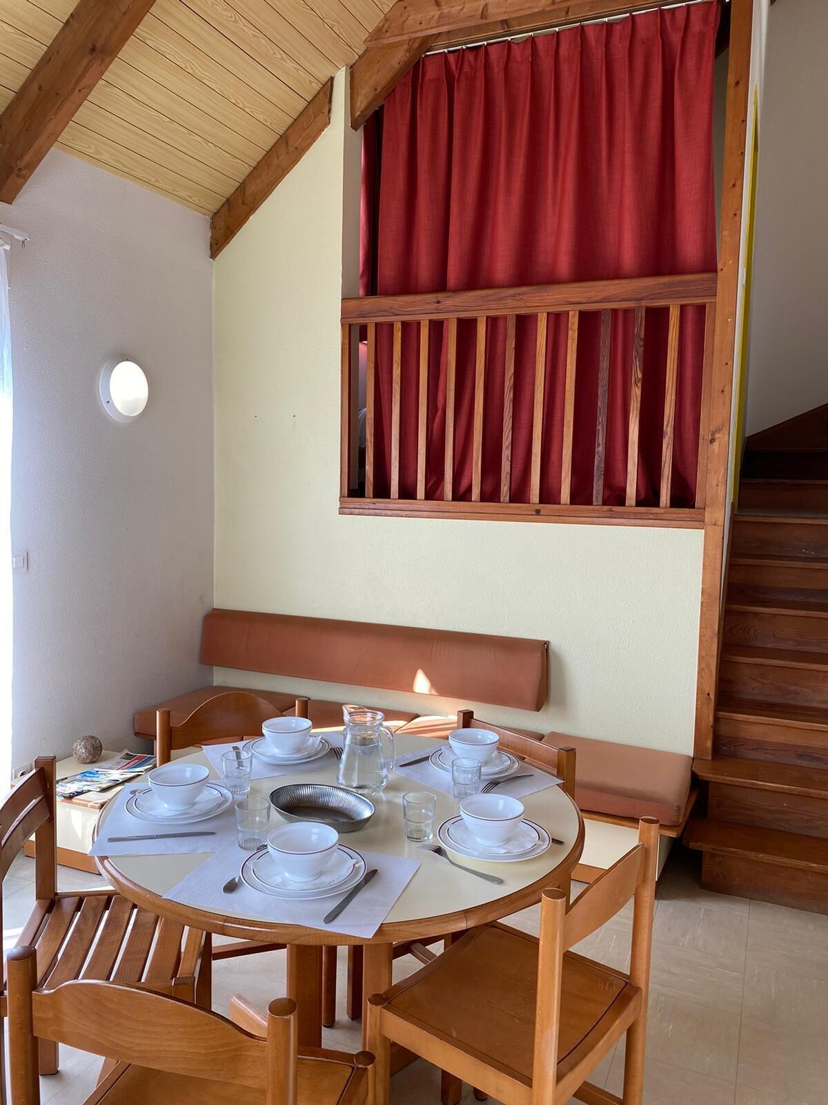 Zona pranzo con tavolo rotondo imbandito per quattro persone, sedie in legno, una panca e una tenda rossa sopra una finestra.
