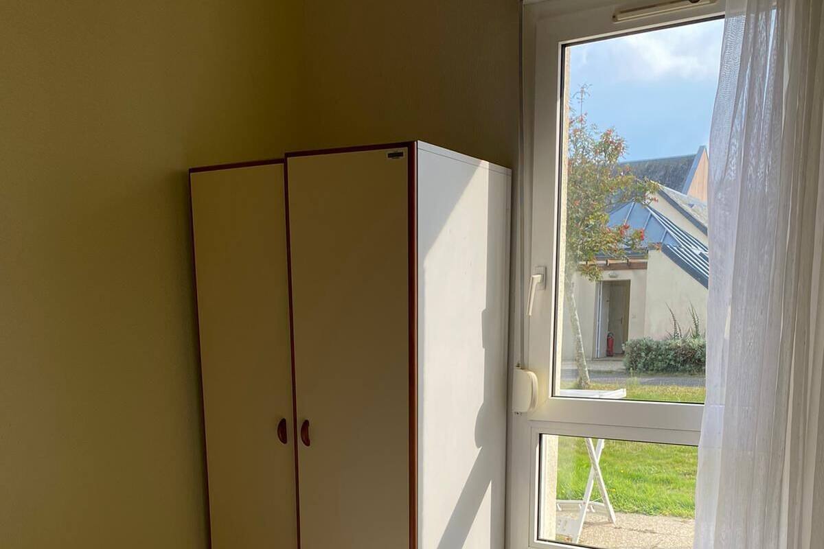 A bedroom with a bed, two towels, a wardrobe, and a window with a view of a building and greenery.