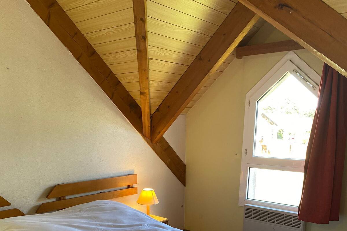 A bedroom with a large bed, wooden beams, a window with curtains, and a bedside lamp.