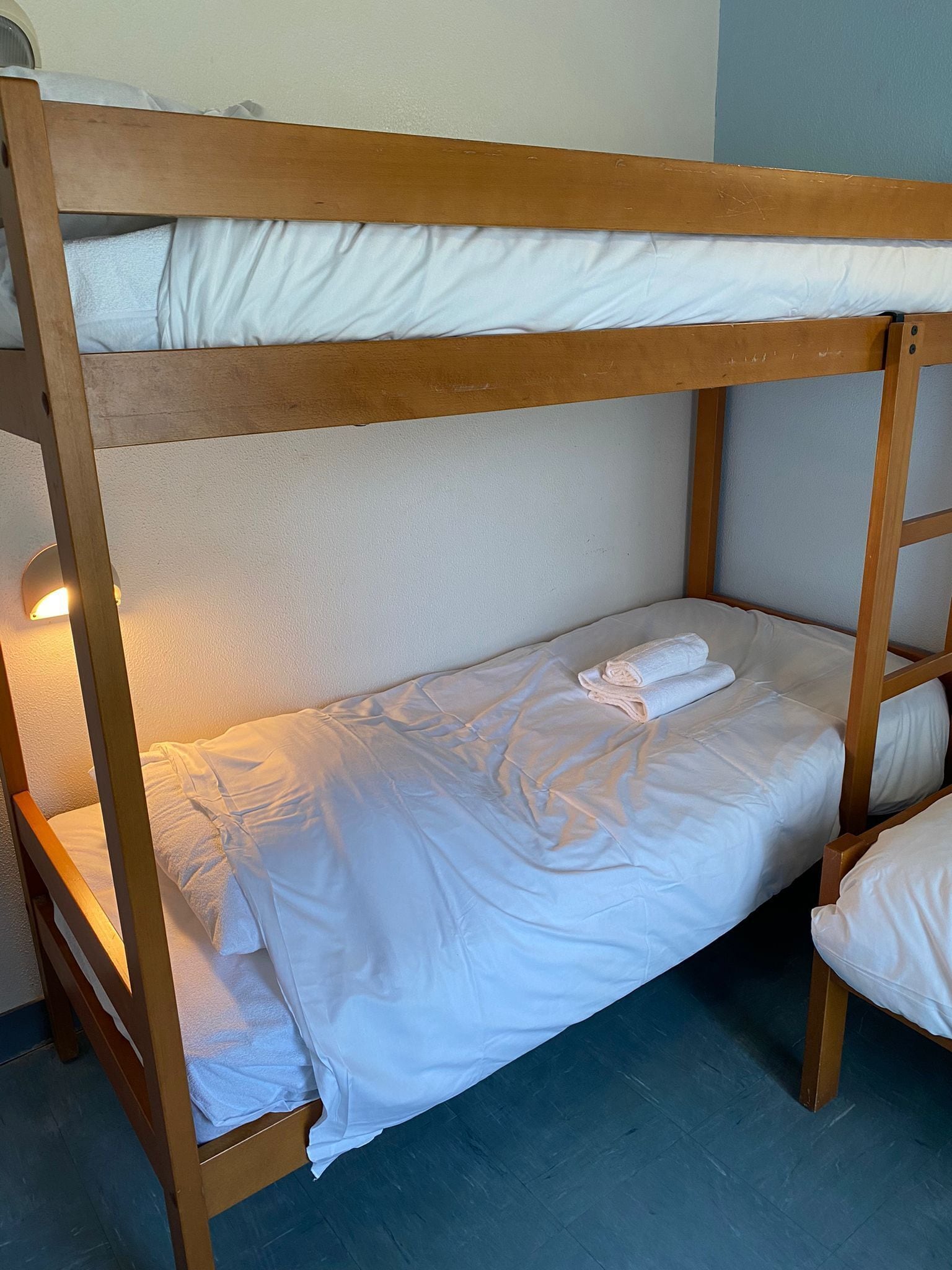 A bunk bed with white bedding and pillows, wooden frame, and a wall-mounted light.