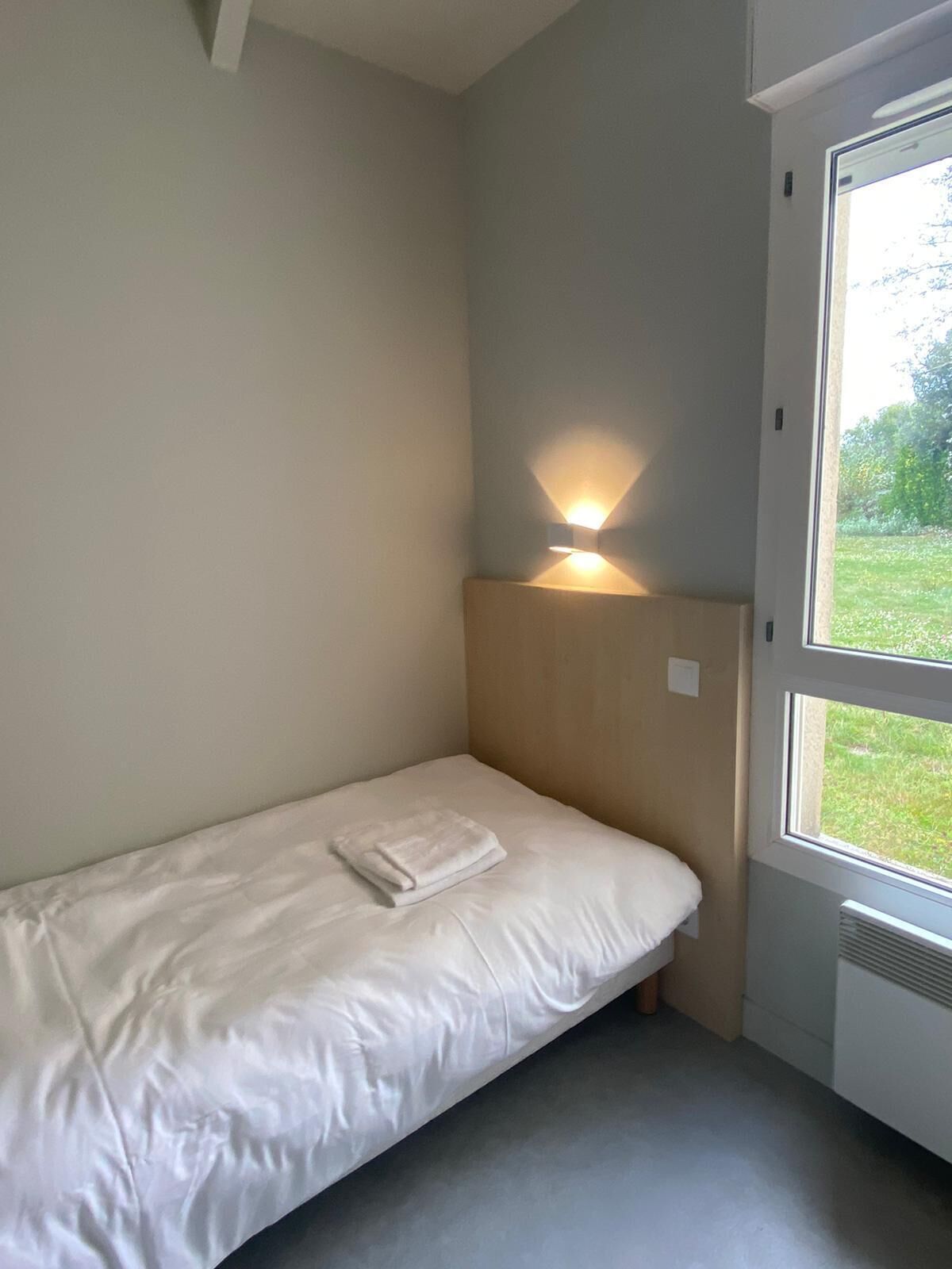A small, single-bed room with a bedside table, a wall-mounted light, and a window with a view of greenery outside.