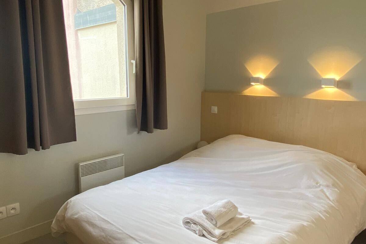 A neatly made bed with a rolled towel, a window with curtains, and two wall-mounted lights.