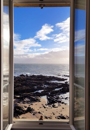 En vy genom ett fönster som visar en stenstrand med vågor, en klar himmel och en person som står längst ner på stranden.